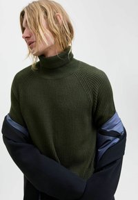 Dunkelgrüner, gerippter Rollkragenpullover mit hohem Kragen, der Raglanärmel hat. Akzentuiert mit einer navyblauen Außenschicht, die teilweise sichtbar ist.