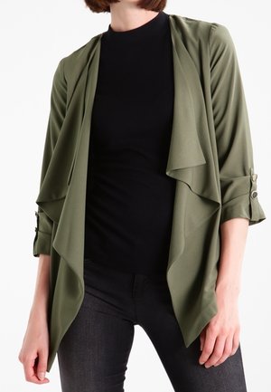 Vrouw die een olijfgroene open voorkant gedrapeerde jas draagt over een zwart aansluitend top en donkergrijze broek, staand tegen een witte achtergrond.