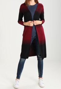 Lång kofta i gradient från burgundy till svart. Tillverkad av mjukt stickat material, med öppen framsida och långa ärmar, kombinerad med en blå topp och jeans.