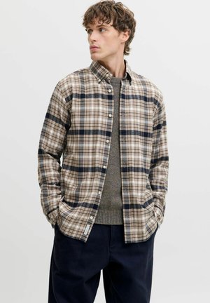 Young man wearing a beige and black plaid button-up shirt over a gray sweater with hands in pockets, looking to the side against a plain background.