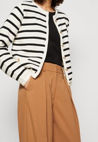 Cardigan a righe in nero e crema, con scollatura rotonda, bottoni dorati e texture a coste, indossato sopra un top nero e pantaloni beige.