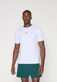 Valge lühikeste varrukate ja sportliku lõikega T-särk ümmarguse kaelusega ja EA7 logoga rinnas, koos roheliste lühikesed pükstega. Sile tekstuur ja sobiv disain.