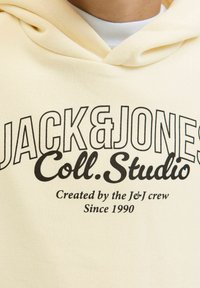 Pogled na kapuco iz blizu, bele barve, s črnim napisom "JACK&JONES Coll. Studio Created by the J&J crew Since 1990" na sprednji strani.