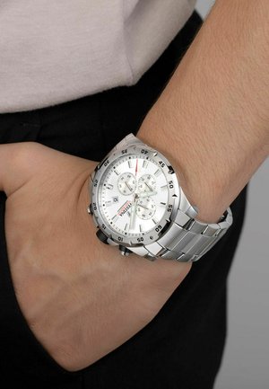Montre en acier inoxydable avec un cadran blanc, trois sous-cadrans, accents noirs et un bracelet métallique, portée sur un poignet humain.