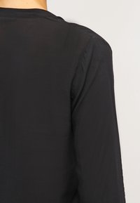 Back view of a person wearing a plain black long-sleeve shirt against a light neutral background.