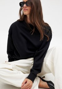 Sudadera negra de gran tamaño con mangas largas, combinada con pantalones de pierna ancha en color blanco sucio. La modelo lleva puestas grandes gafas de sol negras, sentada contra un fondo neutro.