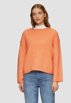 Pull orange surdimensionné avec une texture ribbée, des poignets larges et un col rond, associé à un jean bleu taille haute.