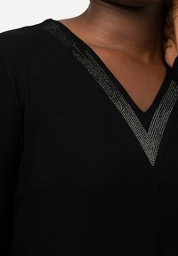 Czarna bluzka z dekoltem w kształcie litery V, z teksturowanym dekoltem zdobionym małymi srebrnymi ćwiekami, wykonana z miękkiego materiału o gładkim wykończeniu.