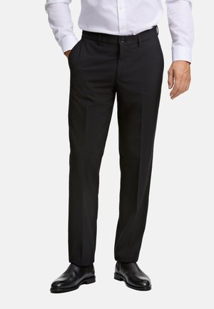 Zwarte pantalon met een rechtlopende snit, gemaakt van een gladde stof, met een knoopsluiting en zijzakken. Draagt een paar gepolijste zwarte schoenen.