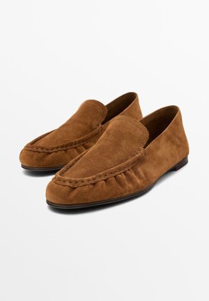 Een paar bruine suède loafers met afgeronde neuzen, gestikte details en lage zwarte zolen, afgebeeld tegen een effen witte achtergrond.