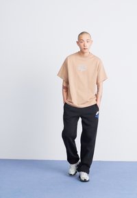 Bež majica s bijelim Nike logotipom, uparena s crnim trenirkama koje imaju detalj s logotipom. Model nosi višebojnu sportsku obuću.