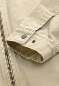 Beige jacket fabric with a textured, woven design; features a brown zipper and metallic snap buttons on the cuff.