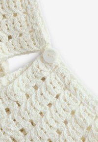 Tissu blanc en crochet avec un motif en diamant, doté d'un grand bouton en plastique à l'épaule. Design doux, texturé et léger.