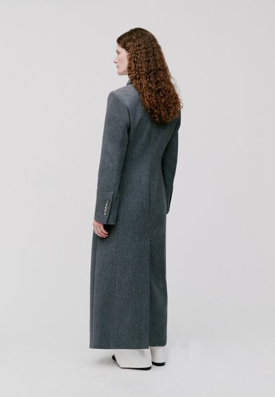 Lungo cappotto di lana grigia con una silhouette su misura, rever a punta e dettagli con bottoni sui polsini, dotato di una spacco sul retro per facilitare il movimento.