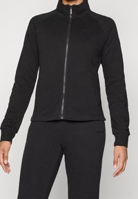 Svart zip-up jacka i jersey tyg med hög krage och raglanärmar. Designen inkluderar en diskret varumärkeslogotyp.