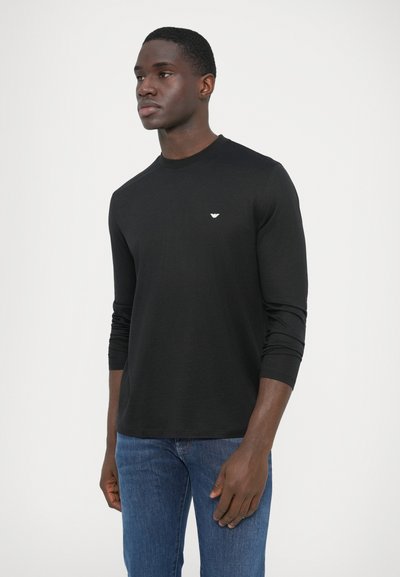 Camiseta negra de manga larga hecha de tela suave con cuello redondo. Presenta un pequeño logo blanco en el pecho. Combinada con vaqueros azules.