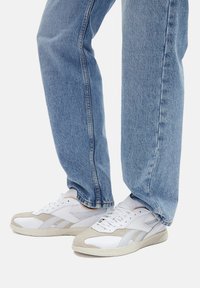 Balti žemakulniai sportbačiai su šviesiai pilkais oda akcentais, perforuotais detalėmis ir tekstūruotais guminiais padais; derinami su šviesiai mėlynos spalvos džinsais.