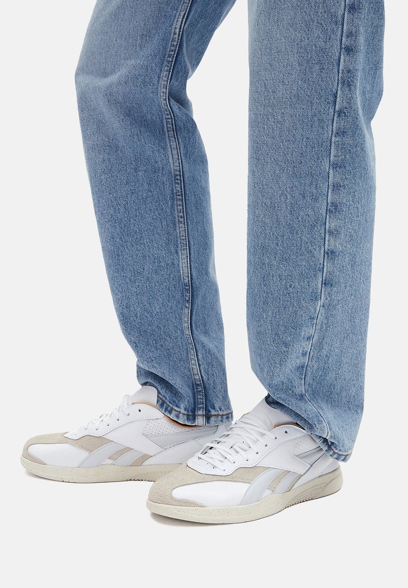 Balti žemakulniai sportbačiai su šviesiai pilkais oda akcentais, perforuotais detalėmis ir tekstūruotais guminiais padais; derinami su šviesiai mėlynos spalvos džinsais.