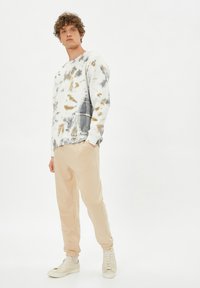 Sudadera blanca con diseño de tie-dye en tonos gris y marrón, combinada con pantalones de chándal beige y zapatillas crema. Tejido suave, corte relajado, diseño minimalista.