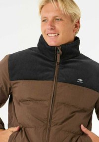 Bruine pufferjack met een zwart corduroy kraag, voorzien van een rits en zijzakken. Gestructureerde stof met een gladde afwerking.