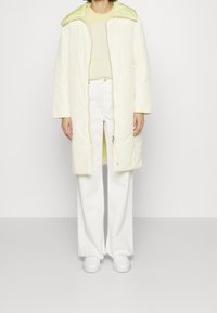 Mujer vestida con un abrigo largo acolchado de color amarillo claro sobre un suéter amarillo pálido, pantalones blancos de cintura alta y zapatillas blancas, de pie contra una pared sencilla.