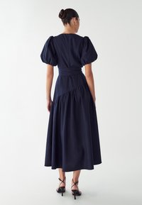 Navyblauwe jurk met pofmouwen, een gedefinieerde taille, een rucheszoom en een ritssluiting aan de achterkant. Gecombineerd met zwarte strappy hoge hakken.