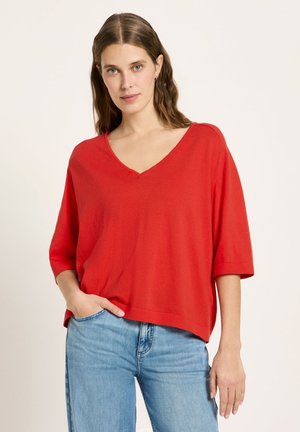 Junge Frau mit langen braunen Haaren trägt ein rotes, locker sitzendes V-Ausschnitt-Oberteil und hellblaue Jeans, steht vor einem schlichten hellen Hintergrund.