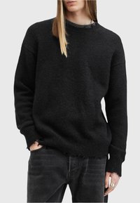 Persoană îmbrăcată cu un pulover negru uzat din tricot, purtând o lanț, blugi negri, cu o mână în buzunar și un inel argintiu pe cealaltă mână.