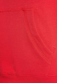 Zbliżenie na czerwoną tkaninę bluzy z kapturem, z kieszenią kangura i widocznymi detalami przeszyć. Miękka faktura z gładkim wykończeniem.