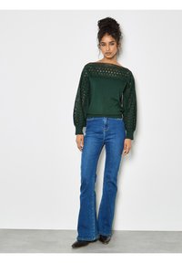 Suéter verde oscuro con detalles de encaje en las mangas y el escote, combinado con jeans de campana azules y botas negras sobre un fondo claro.