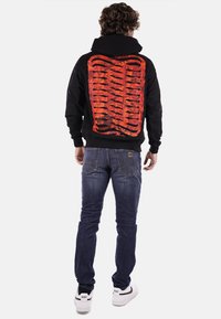 Felpa nera con un audace design grafico rosso che somiglia a delle costole sulla schiena. Abbinata a un paio di jeans blu e scarpe da tennis bianche.