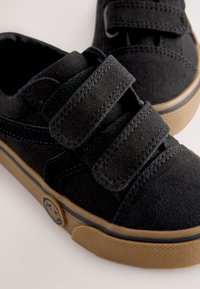 Melnas auduma auduma kurpes ar divām Velcro siksnām, gumijas zolīšu un apaļu logotipu sānos. Teksturēts audums un kontrastējošas šūšanas detaļas.