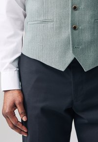 Gilet vert clair texturé à trois boutons, associé à un pantalon foncé. Les poignets de la chemise blanche sont visibles, mettant en valeur un design formel classique.