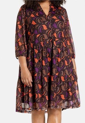 Nainen, jolla on polvipituinen mekko, jossa on kolme neljänneksen hihaa ja abstrakti oranssin, purppuran ja ruskean pyörrekuvio.