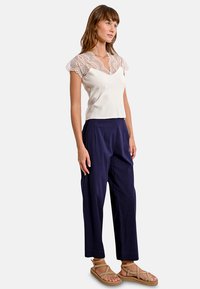 Maglietta beige chiaro con maniche in pizzo, abbinata a pantaloni blu navy larghi e sandali color nocciola. Tessuto morbido con dettagli in pizzo elaborato.