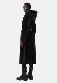 Marineblåt faux fur-jakke med hætte, bælte i taljen og sidelommer med lynlås. Modellen har sorte combat-støvler og et rødt ærme på under Jakken.