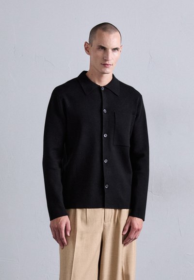 Un modello maschile si appoggia a un muro grigio indossando un cardigan nero con bottoni e tasca sul petto, abbinato a pantaloni beige a pieghe con gamba larga.