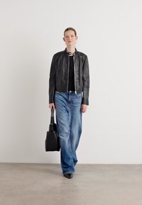 Črna usnjena jakna, črn top in svetlo modri široki hlače. Drži črno torbo. Enostavna ozadje, osredotočeno na oblačila.