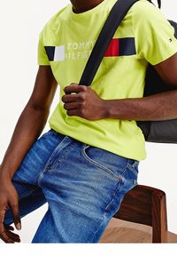 Camiseta de Tommy Hilfiger en verde neón con logo y bloques de color en rojo, azul y blanco. Combinada con vaqueros de mezclilla azules y una mochila negra.