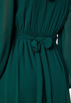 Abito verde fluido con vita arricciata e fiocco. Realizzato in tessuto leggero, ha una consistenza morbida e dettagli plissettati.
