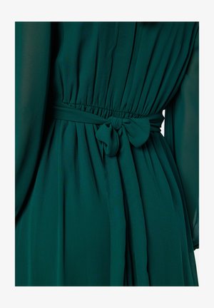 Abito verde fluido con vita arricciata e fiocco. Realizzato in tessuto leggero, ha una consistenza morbida e dettagli plissettati.