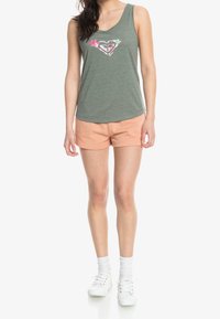 Mujer vestida con una camiseta sin mangas de logo floral verde, pantalones cortos melocotón, calcetines blancos y zapatillas blancas de pie contra un fondo blanco.