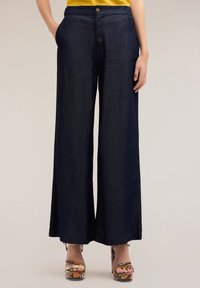 Donkerblauwe wijde broek met een gladde textuur, voorzien van een knoopsluiting en zijzakken, gecombineerd met meerkleurige platform sandalen.
