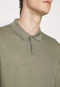 Gros plan sur une personne portant un polo en maille texturée vert olive clair, avec une patte courte à boutons et un col replié.