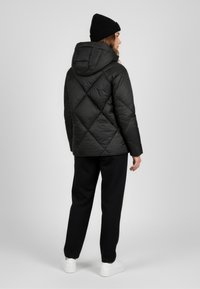 Giubbotto imbottito nero con trapuntatura a diamante e cappuccio, abbinato a pantaloni neri e sneakers bianche. Il tessuto appare lucido e leggero.