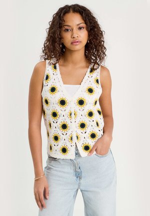 Młoda kobieta o kręconych włosach, ubrana w białą szydełkowaną kamizelkę z wzorami słoneczników na białej bluzce i jasnoniebieskich dżinsach.