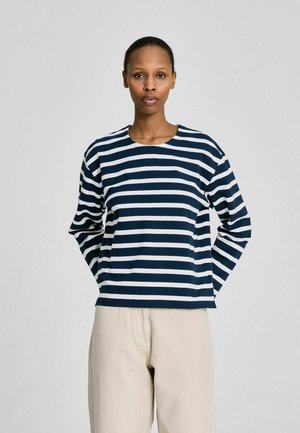 Naine, kes kannab tumesinist ja valget horisontaaltriibulist pika varrukaga särki ning beeže pükse, seisab käed selja taga.