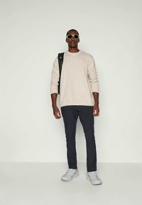 Sweatshirt bege claro com decote redondo, calças azul marinho escuras e ténis cinzentos. Os acessórios incluem óculos de sol brancos e uma mochila preta.