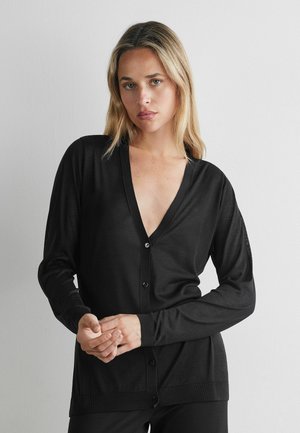 Junge Frau mit blondem Haar, die eine schwarze V-Ausschnitt-Knopfstrickjacke und schwarze Hose trägt, steht vor einer schlichten grauen Wand.