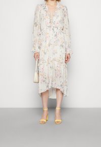 Robe fleurie à manches longues et col à volants, en tissu léger orné de fleurs pastel ; accompagnée de sandales à talons jaunes et d'un sac à main.